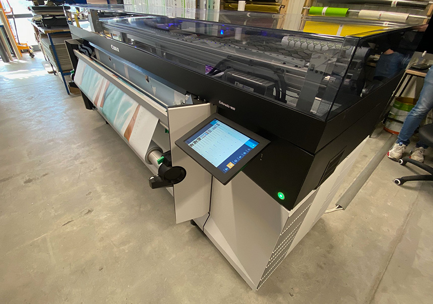 Impression d’un papier peint rayé sur une imprimante Canon Colorado dans l’atelier Manufacture Arssans, avec différents modèles décoratifs exposés au mur.